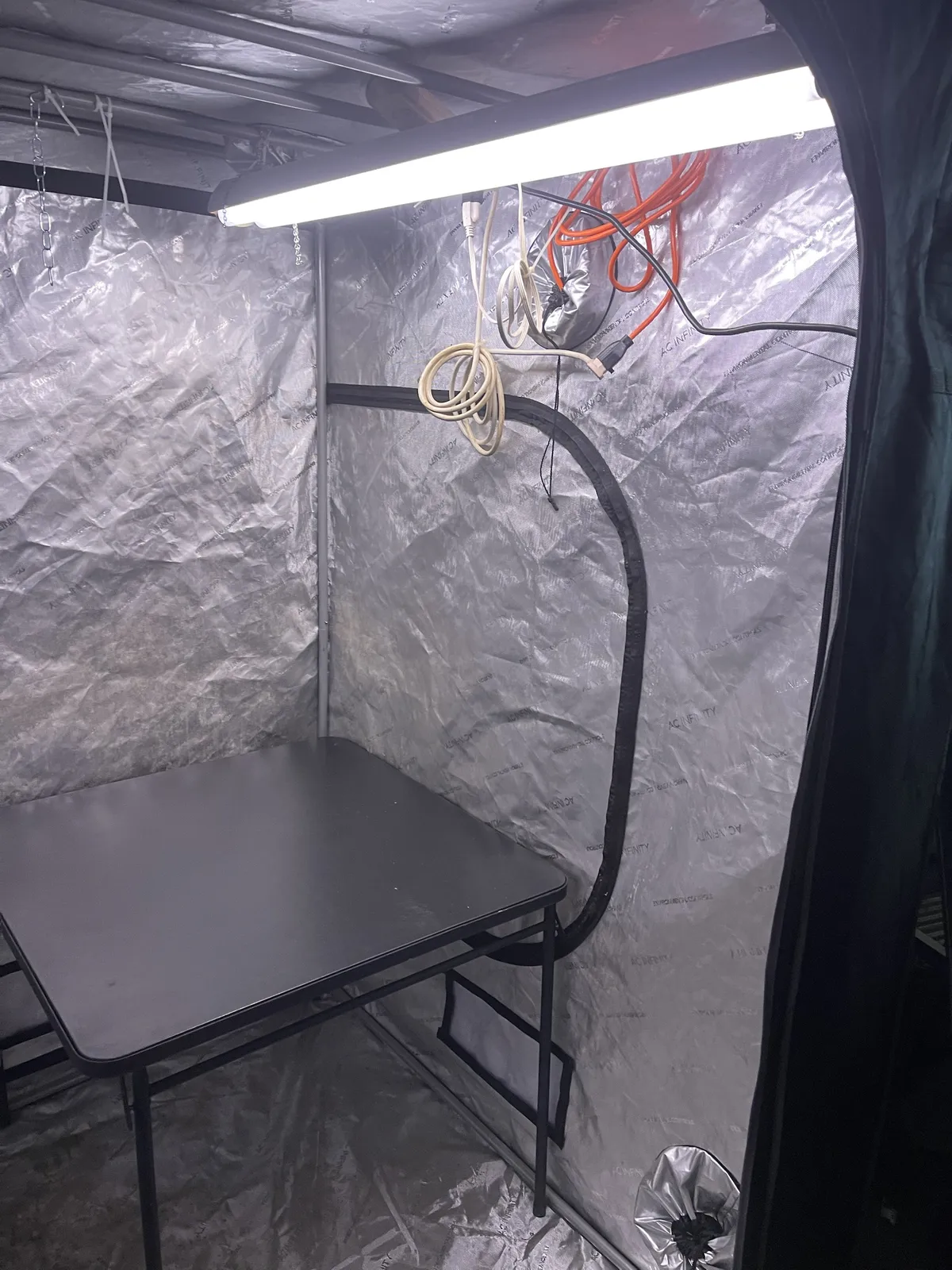 Inside the tent with the light on: a black folding table, the hanging coiled cords, and the silver cinch-top duct ports on the wall and in the floor corner.