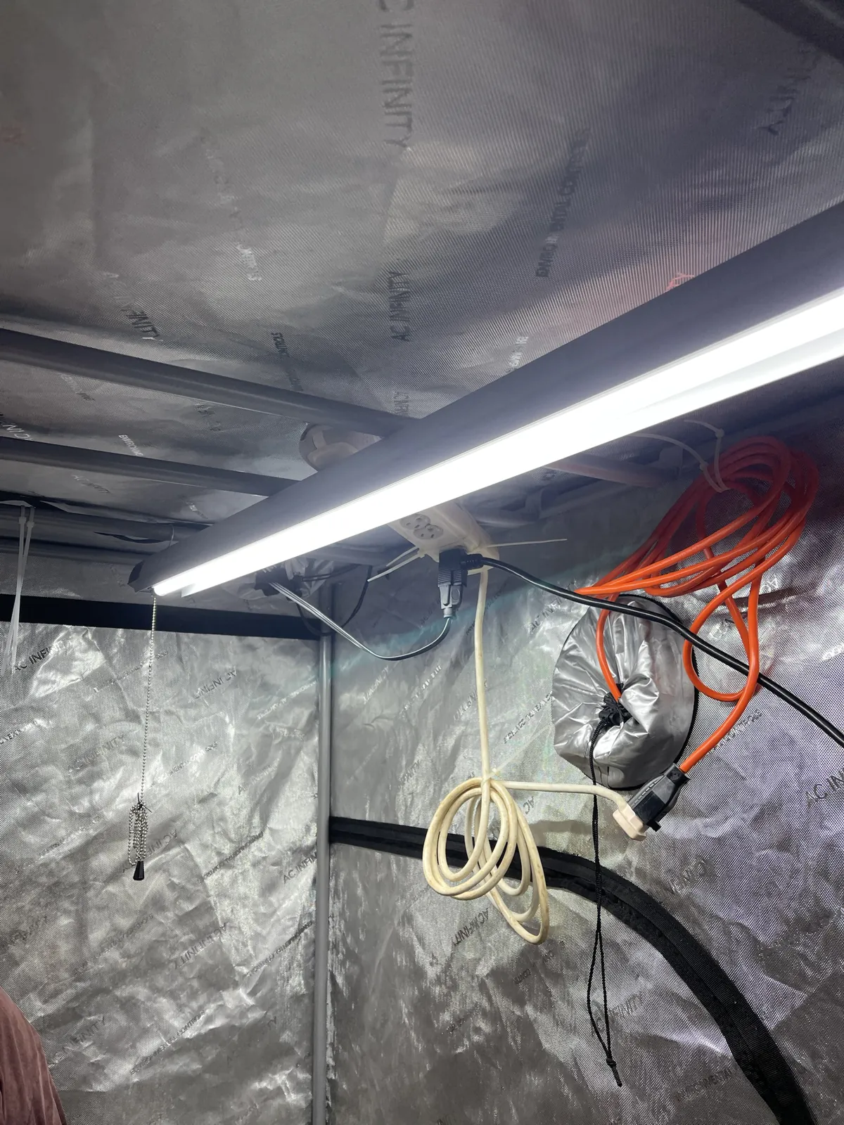 The new LED light on, running along the tent's top frame, with the surge protector zip-tied nearby, the orange primary power cord coiled on the wall, excess length of the surge protector's white cord looped below, and a bead pull chain hanging at left.
