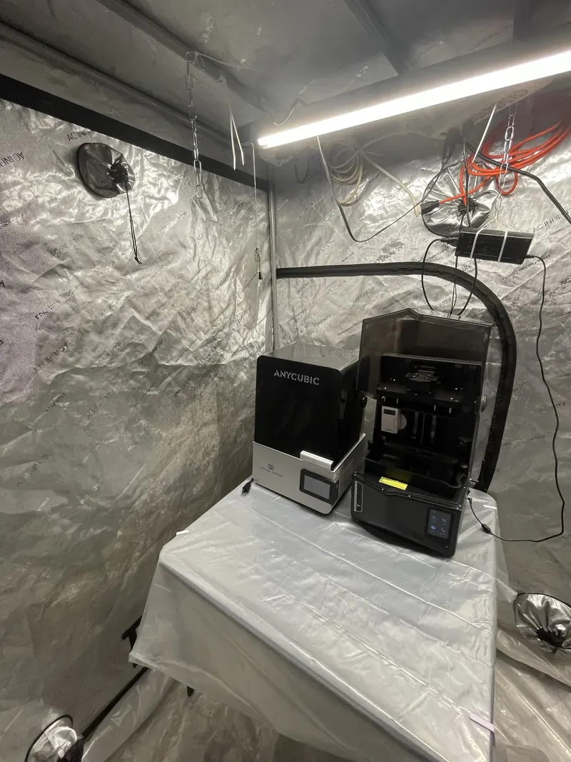 The Anycubic Photon P1 and Elegoo Saturn 4 Ultra on a plastic-covered table inside the garage tent; the Saturn hood is open and its vat is empty.